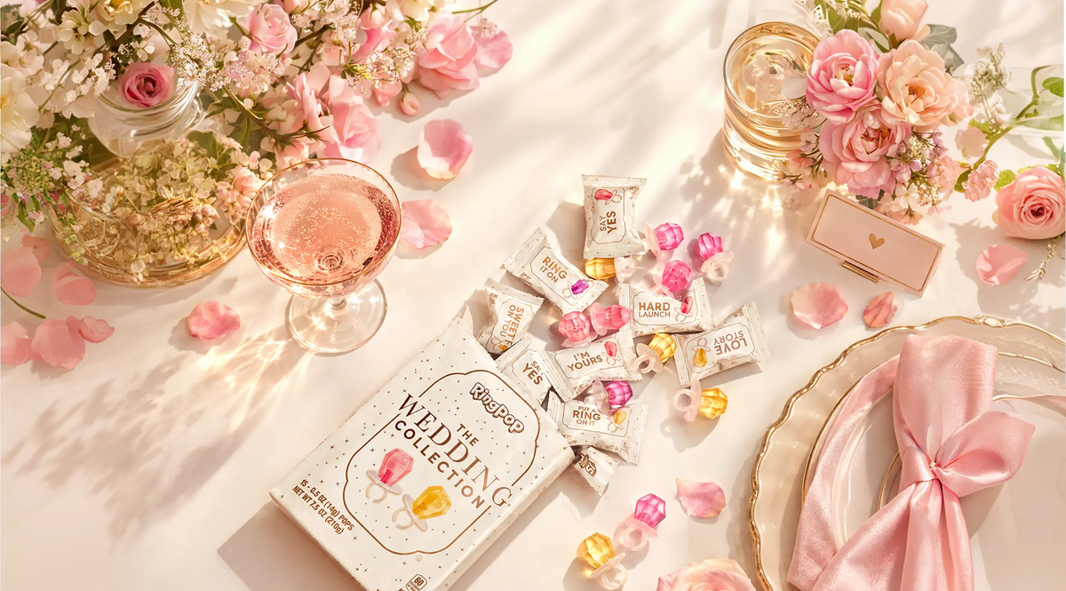 A styled flat-lay of a box of Wedding The Collection soft gummy candies, surrounded by pink rose petals, a pink champagne flute, and elegant tableware on a warm, sunlit cream background.