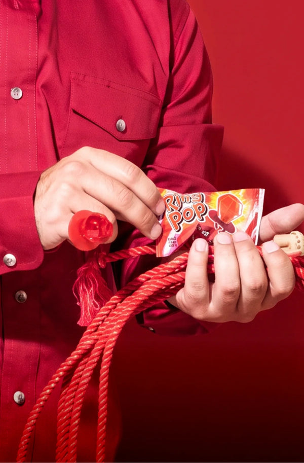 A person in a red button-up shirt holds a small bag of Ring Pop® Colorfest  candy and a red rope against a solid red background.