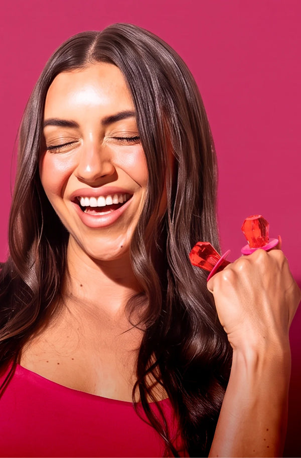 A smiling woman with long dark brown hair holds up two small red rose-shaped rings on her fingers against a solid pink background.