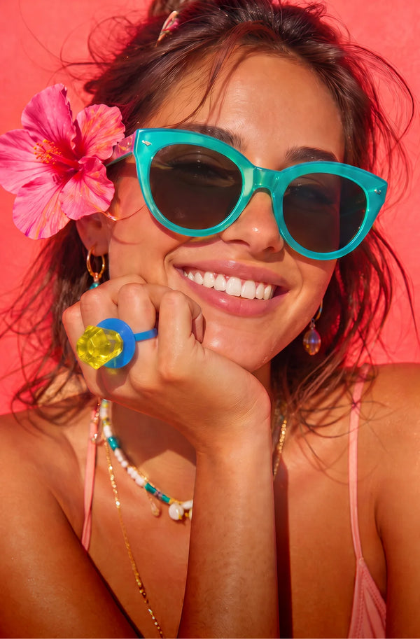 A smiling woman wearing bright turquoise cat-eye sunglasses with dark lenses, posed against a vivid pink background. She has a large pink hibiscus flower in her hair, a colorful Ring Pop® candy on one finger, and layered necklaces, making this a lifestyle shot.