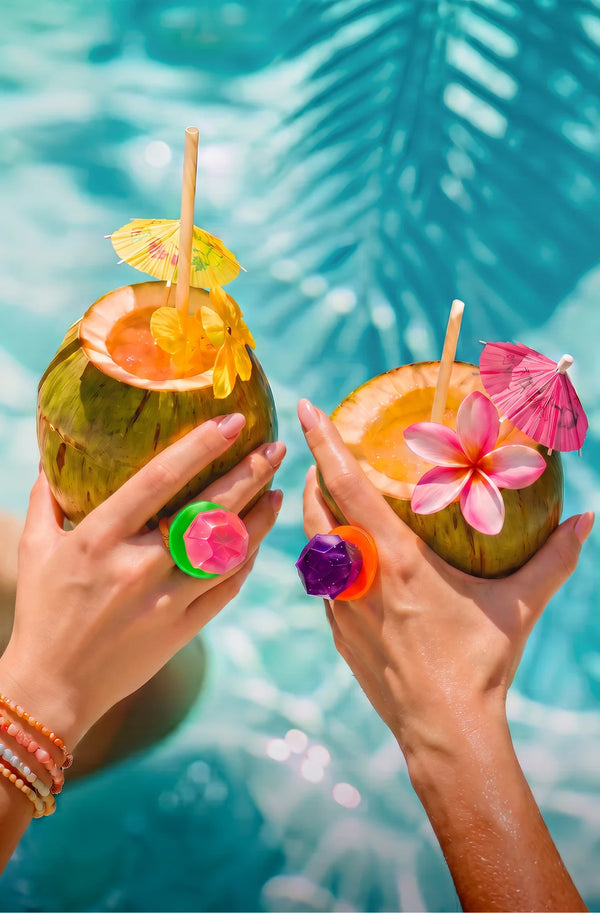 Two hands holding coconut drinks with umbrellas and flowers, set against a bright blue swimming pool background with palm leaves.