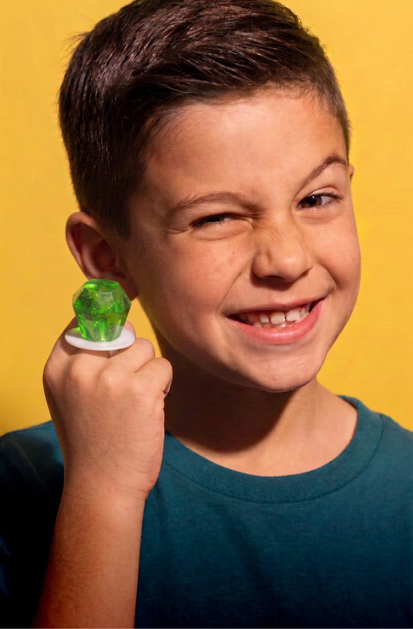 A young boy with short brown hair smiles while wearing a Ring Pop® candy on a white ring shaped like a stylized ear, shown against a solid yellow background.
