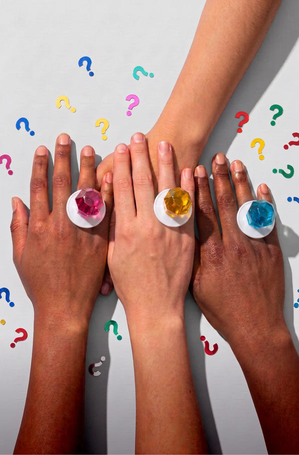Four hands stacked together wearing oversized white rings with colorful Ring Pop® candies in pink, yellow, blue, and green, set against a light gray background with scattered question marks.