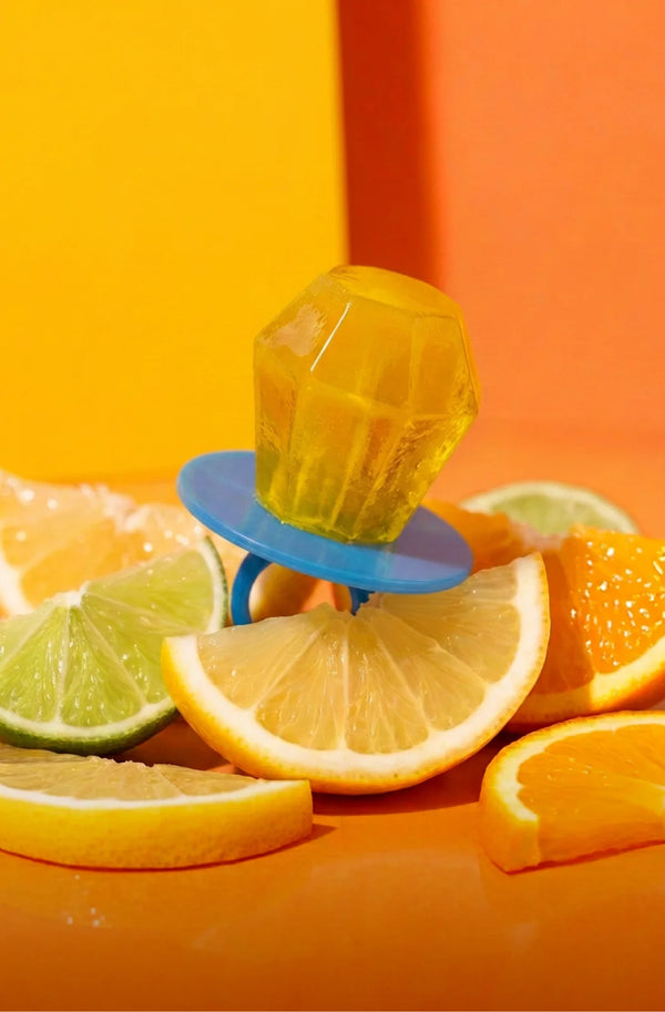 Ring Pop® candy, resting on a blue circular stand, surrounded by citrus slices including orange, lemon, and lime against a bright orange and yellow studio background.