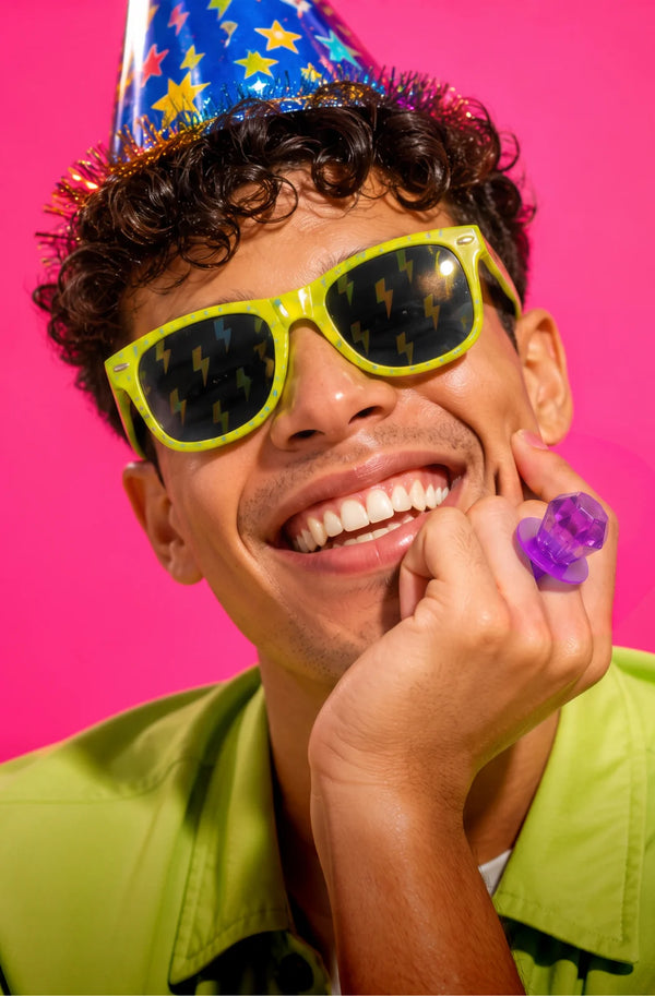 A smiling man wearing a blue party hat with yellow stars, neon yellow sunglasses with lightning-bolt lenses, and a bright lime green shirt, posed against a solid hot pink background.