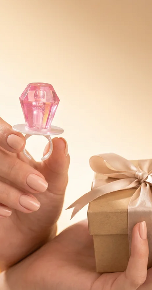 A hand holds a novelty ring shaped like a faceted Ring Pop®  on a white ring stand, with another hand holding a small brown gift box tied with a satin ribbon in the background, against a warm beige backdrop.